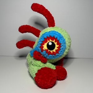 Può includere: Un giocattolo di peluche all'uncinetto con un corpo verde, braccia e gambe rosse e un occhio blu e giallo. Il giocattolo ha un design fantasioso e colorato.