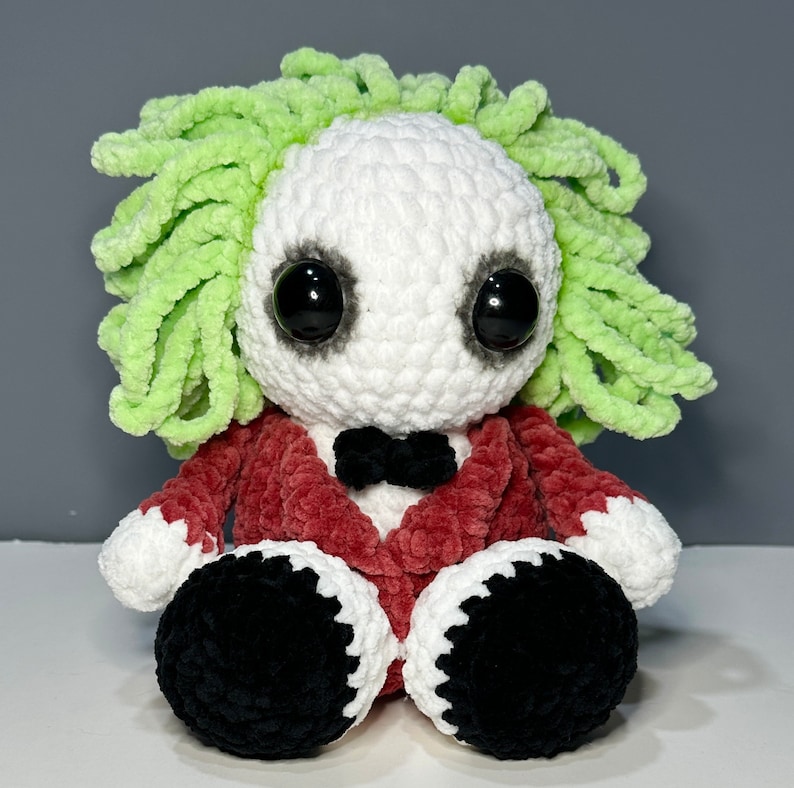 May include: A crocheted plush toy with a white face, black eyes, and a red jacket. The toy has green hair and black shoes.