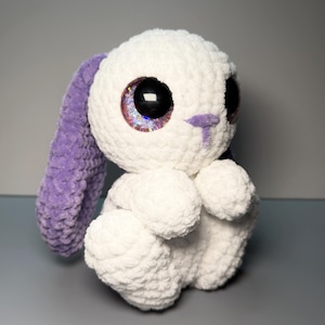 May include: A handmade white crochet bunny with large, glittery eyes and purple accents on the ears and nose. The plush toy is soft and cuddly, perfect for a child's room or as a decorative item. The bunny is sitting with its arms crossed.