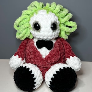 May include: A crocheted plush toy with a green hair-like top, a white face with black eyes, a red jacket, and black shoes.