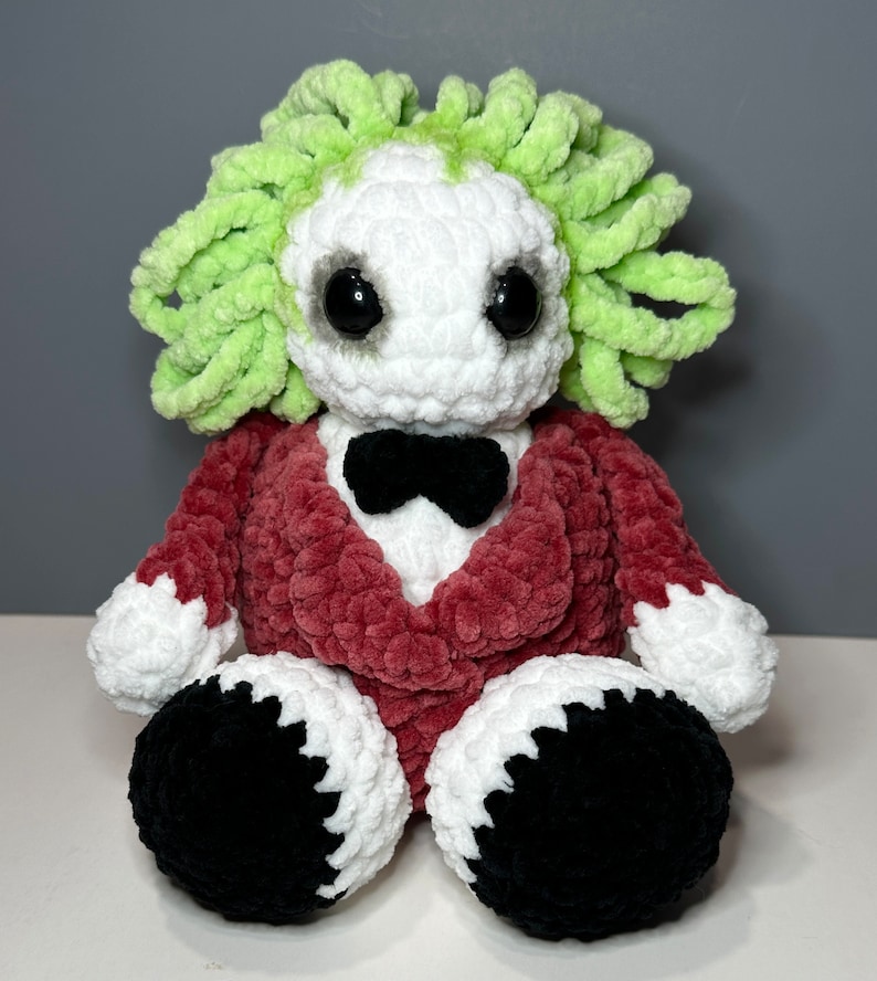 May include: A crocheted plush toy with a green hair-like top, a white face with black eyes, a red jacket, and black shoes.