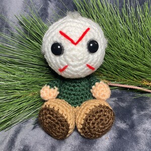 Puede incluir: Un juguete de peluche tejido a crochet de un personaje con cabeza blanca, camisa verde y zapatos marrones. El personaje tiene una X roja dibujada en la cara y ojos negros.