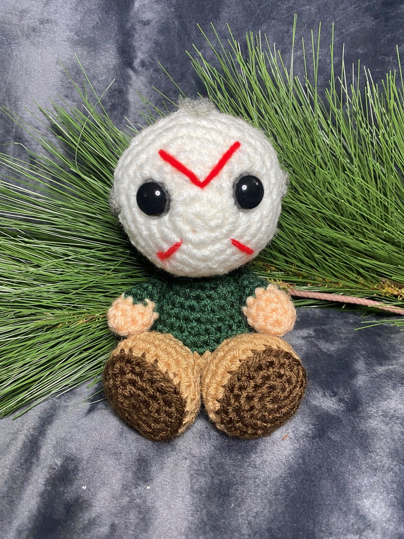 Puede incluir: Un juguete de peluche tejido a crochet de un personaje con cabeza blanca, camisa verde y zapatos marrones. El personaje tiene una X roja dibujada en la cara y ojos negros.