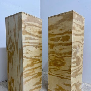 May include: Two rectangular wooden pedestals with a natural wood grain finish. The pedestals are tall and box-shaped, suitable for displaying items. The wood has a light brown color with darker grain patterns.
