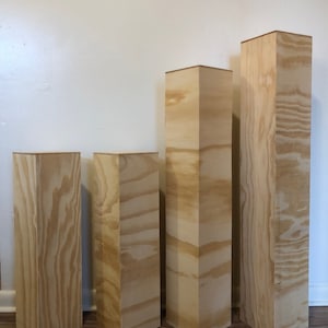 May include: Four unfinished wooden display pedestals of varying heights. The pedestals are rectangular with a natural wood grain finish. They are arranged in a row, with the tallest one on the right and the shortest on the left. The pedestals are suitable for displaying art or products.