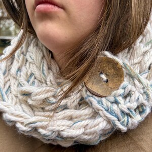 May include: A chunky knit cowl in cream with blue and yellow accents. The cowl features a large wooden button closure. The thick yarn creates a textured, warm appearance. This neck warmer is a cozy accessory for cold weather.