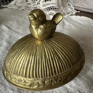 Puede incluir: Una caja de baratijas de latón vintage con una tapa en forma de pájaro. La tapa tiene forma de cúpula con líneas verticales y un borde floral decorativo. El pájaro está posado encima, mirando hacia adelante. La caja probablemente se usa para guardar objetos pequeños.