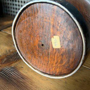 May include: Close-up of the bottom of a wooden barrel. The barrel has a dark brown finish with a light-colored rim and black metal bands. A small, rectangular sticker with the number "1064" is affixed to the wood.