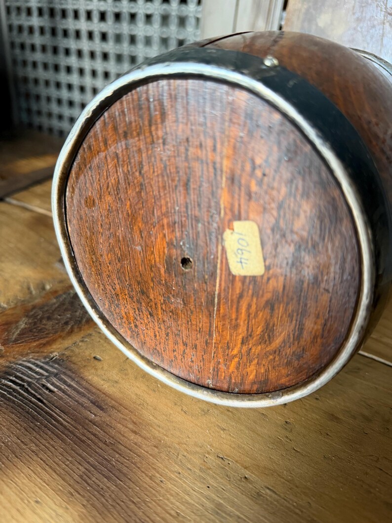May include: Close-up of the bottom of a wooden barrel. The barrel has a dark brown finish with a light-colored rim and black metal bands. A small, rectangular sticker with the number "1064" is affixed to the wood.