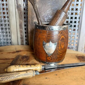 May include: A wooden barrel-shaped utensil holder with a silver rim and shield-shaped emblem. It contains a wooden spoon and rolling pin. Two knives with light-colored handles rest on a wooden surface, suggesting a kitchen or dining theme.