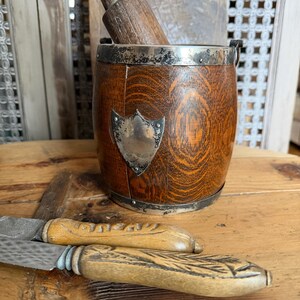 May include: A wooden mortar and pestle set with a silver shield emblem and metal trim. Two antique knives with wooden handles are in the foreground. The items are displayed on a wooden surface.