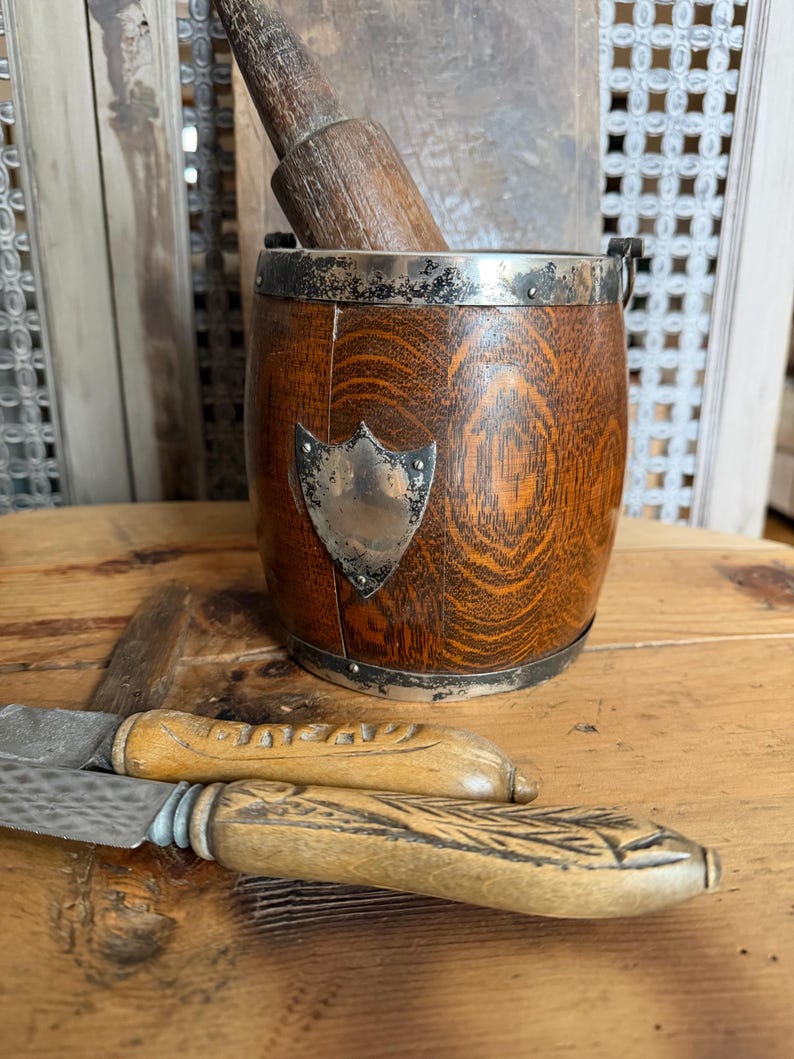 May include: A wooden mortar and pestle set with a silver shield emblem and metal trim. Two antique knives with wooden handles are in the foreground. The items are displayed on a wooden surface.