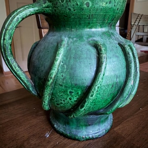 May include: A large, round ceramic pitcher with a vibrant green glaze and a mottled, dark-spotted texture. The vessel features a wide, flared rim, a sturdy side handle, and several raised, curved vertical ribs extending from the base to the shoulder.