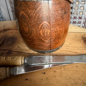 May include: A wooden bucket with a silver rim and handle, alongside two knives with wooden handles. The bucket has a natural wood grain pattern. The knives have silver blades and are resting on a wooden surface.
