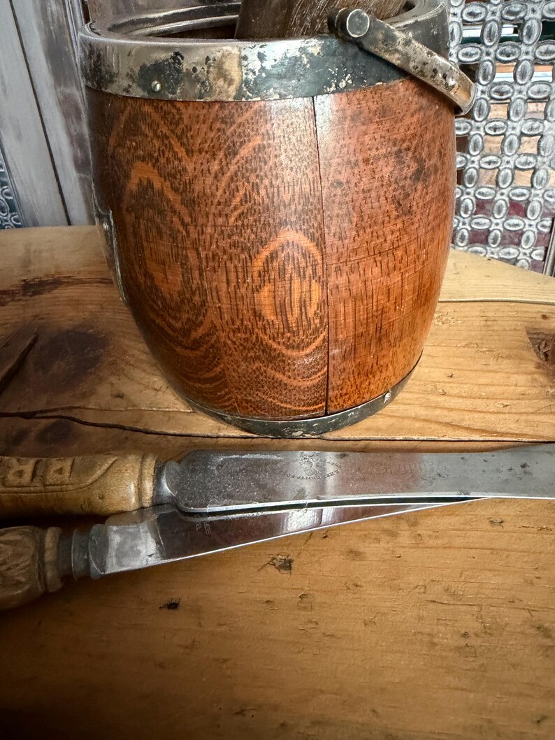 May include: A wooden bucket with a silver rim and handle, alongside two knives with wooden handles. The bucket has a natural wood grain pattern. The knives have silver blades and are resting on a wooden surface.