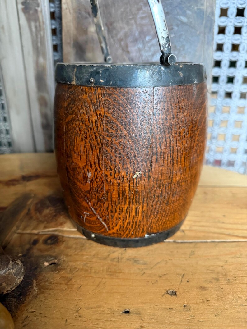 May include: A small, wooden barrel with a metal rim and handle. The barrel has a dark brown, aged appearance with visible wood grain. The metal rim and handle are silver and black. The barrel is likely a decorative item or small container.