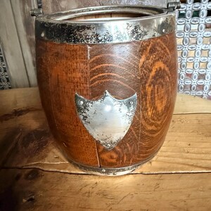 May include: A wooden ice bucket with a silver rim and a shield-shaped emblem. The bucket has a barrel shape with vertical wooden staves. The silver rim and emblem show signs of age. The bucket sits on a wooden surface.