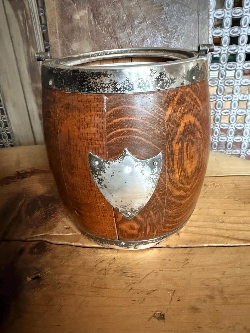 May include: A wooden ice bucket with a silver rim and a shield-shaped emblem. The bucket has a barrel shape with vertical wooden staves. The silver rim and emblem show signs of age. The bucket sits on a wooden surface.