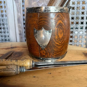 May include: A wooden barrel-shaped container with a silver rim and a silver shield emblem. The container holds wooden tools. A knife with a wooden handle rests on a wooden surface.