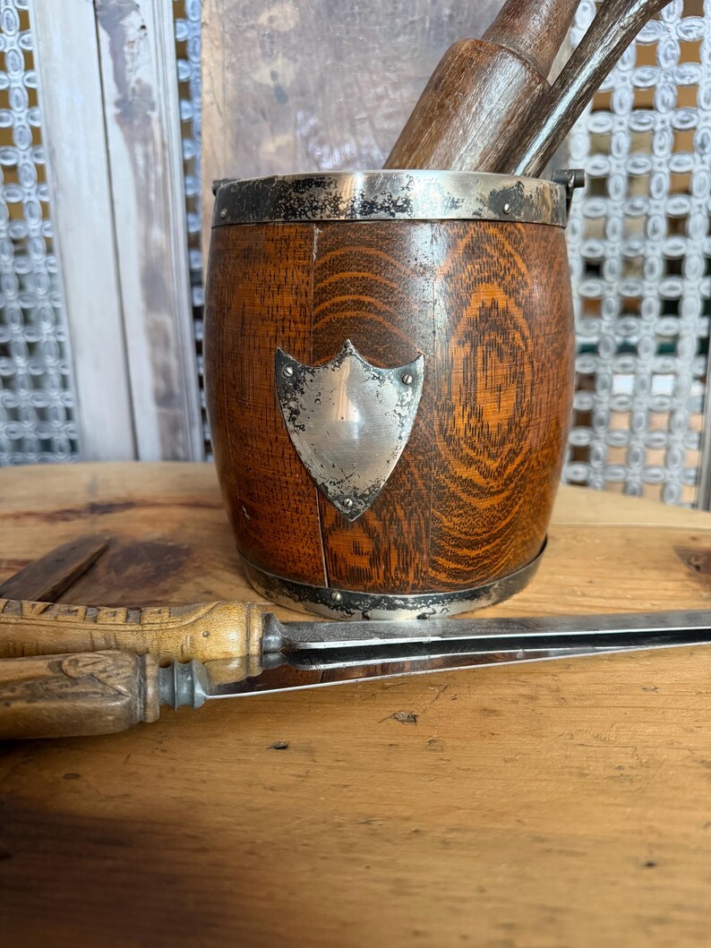 May include: A wooden barrel-shaped container with a silver rim and a silver shield emblem. The container holds wooden tools. A knife with a wooden handle rests on a wooden surface.