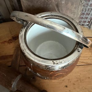 May include: An antique ice bucket with a silver-plated rim and handle. The bucket has a brown, wooden exterior and a white interior. The handle is raised, and the bucket is sitting on a wooden surface. A wooden bread tool is in the foreground.