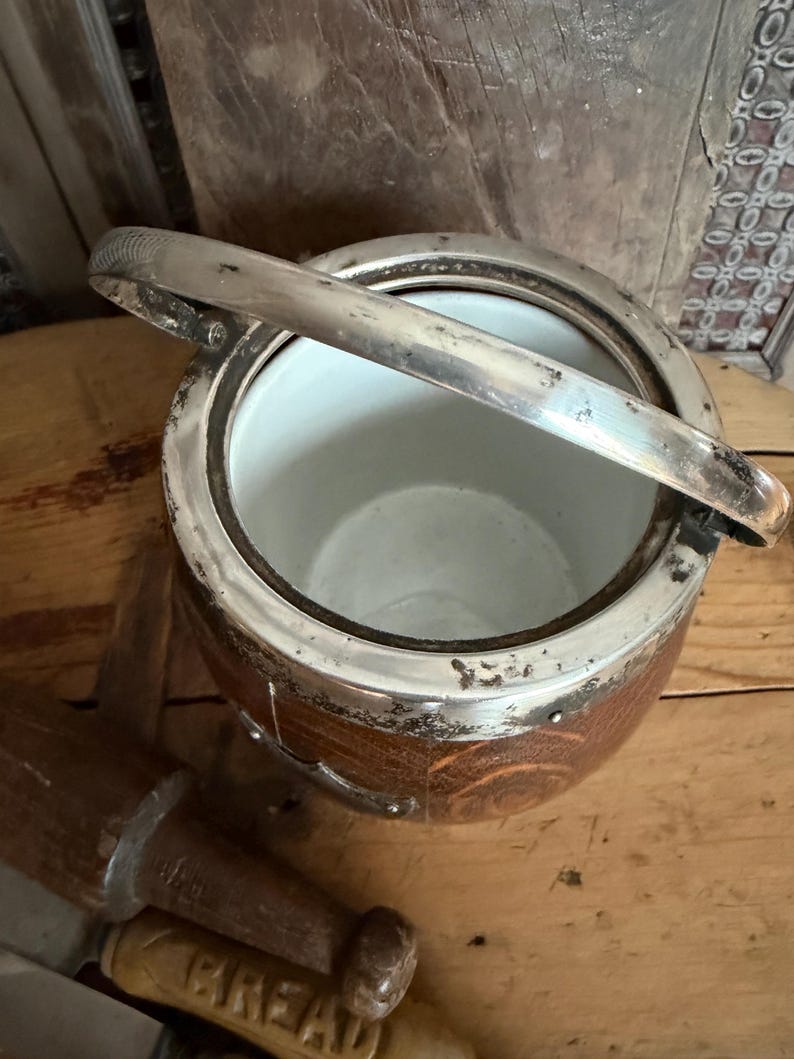 May include: An antique ice bucket with a silver-plated rim and handle. The bucket has a brown, wooden exterior and a white interior. The handle is raised, and the bucket is sitting on a wooden surface. A wooden bread tool is in the foreground.