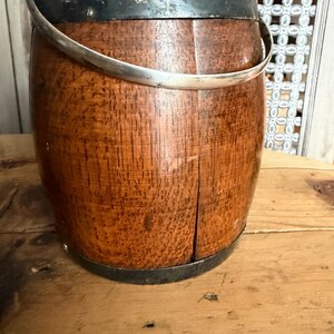 May include: A wooden bucket with a silver handle and metal bands around the top and bottom. The bucket has a rich, reddish-brown color and a visible wood grain texture. The handle is curved and attached to the bucket with metal brackets.