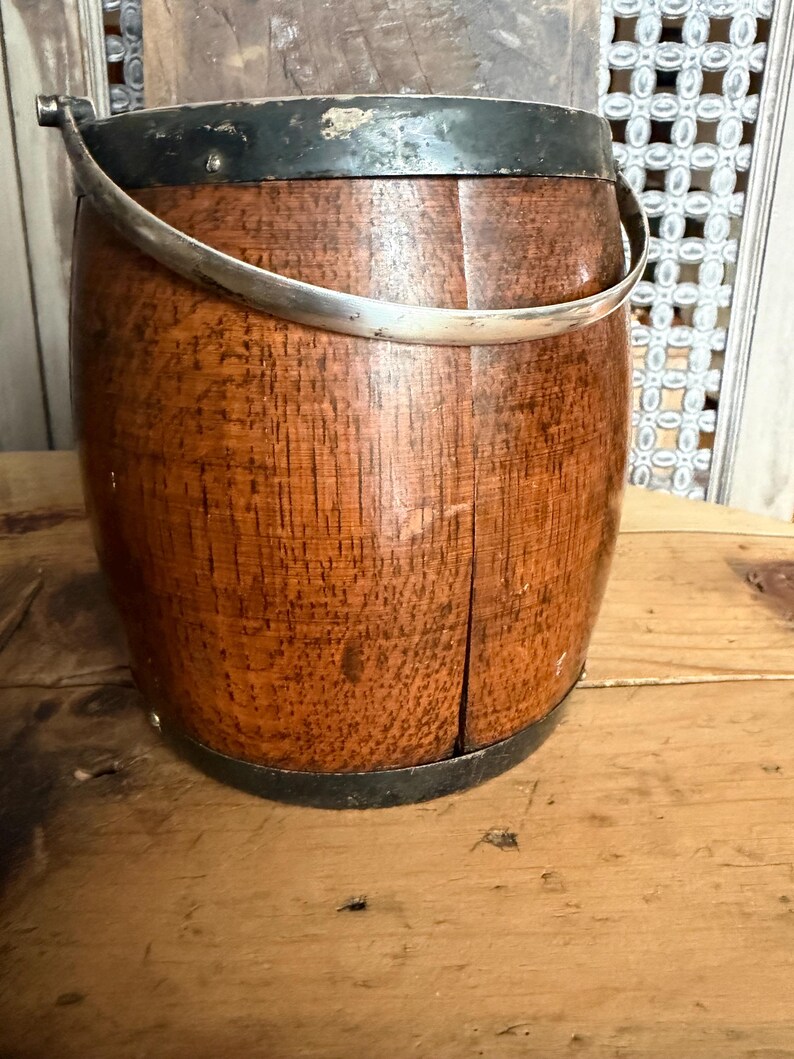 May include: A wooden bucket with a silver handle and metal bands around the top and bottom. The bucket has a rich, reddish-brown color and a visible wood grain texture. The handle is curved and attached to the bucket with metal brackets.