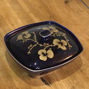 May include: A small, square, cobalt blue ceramic dish with a lid. The dish is decorated with a gold floral design.