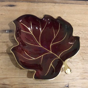 Può includere: Un piatto a forma di foglia di colore bordeaux con accenti dorati. Il piatto è in ceramica e ha una finitura lucida.