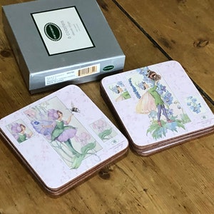 May include: A set of six coasters featuring a fairy design with purple flowers and a bee. The coasters are made of cork and have a pink background. The box for the coasters is silver with the text "6 Coasters" printed on it.
