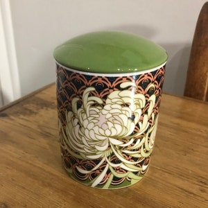 Puede incluir: Un tarro de cerámica con tapa verde y diseño floral. El tarro presenta una flor de crisantemo blanca con hojas verdes y un fondo negro y naranja.