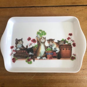 May include: White rectangular serving tray with rounded edges and handles. The tray features a colorful illustration of several kittens among potted plants with red flowers. The kittens are in various poses, adding a playful touch to the design.