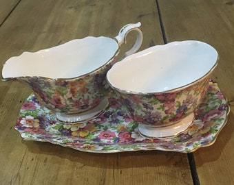Pode incluir: Um conjunto de jarro de creme e açucareiro com padrão floral e uma bandeja combinando. O conjunto é feito de porcelana branca com detalhes dourados e apresenta uma variedade de flores coloridas.
