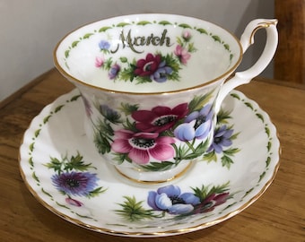 Puede incluir: Una taza de té y un platillo blancos con un diseño floral. La taza tiene un borde dorado y las palabras "March" escritas en ella. El platillo tiene un borde festoneado.