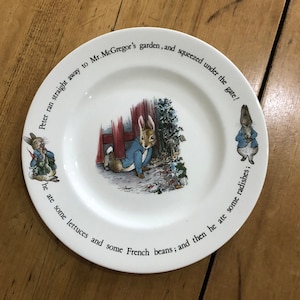 May include: A white ceramic plate with a blue and brown illustration of Peter Rabbit from the children's book by Beatrix Potter. The plate has a border with text that reads "Peter ran straight away to Mr. McGregor's garden, and squeezed under the gate! The ate some lettuces and some French beans; and then he ate some radishes;"
