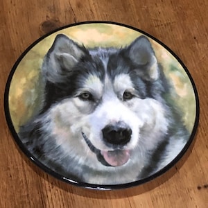 Op de afbeelding: Een keramische plaat met een geschilderd beeld van een witte en grijze Alaskan Malamute-hond met zwarte ogen en een roze tong. De hond kijkt recht naar de kijker met een gelukkige uitdrukking.