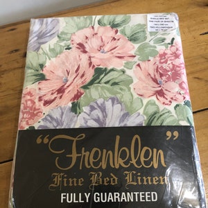 Op de afbeelding: Een pakket Frenklen Fijne Beddengoed, een eenpersoonsbedset van 100% katoen. De verpakking heeft een bloemenpatroon met roze, paarse en groene bloemen op een witte achtergrond. Het pakket bevat een paar lakens van 160 x 230 cm en twee kussenslopen van 50 x 70 cm.