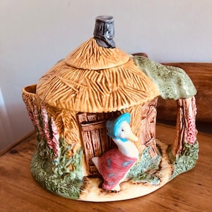 以下が含まれることがあります： 青いボンネットをかぶったガチョウが外に立っている、わらぶき屋根の小屋の形をしたセラミック製のティーポット。ティーポットは、茶色、緑、ピンクのディテールで飾られています。