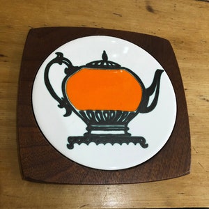 May include: A round ceramic coaster featuring a black and white silhouette of a teapot with an orange interior. The coaster is framed by a dark wood square.