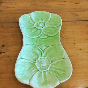 May include: A light green ceramic serving platter with a floral design. The platter is shaped like a flower with embossed petals and a central flower detail. The platter is on a wooden surface.