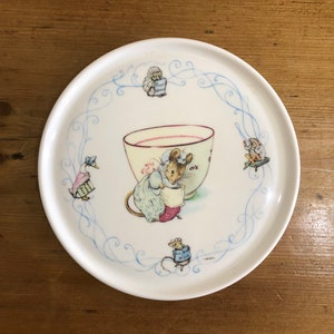 Può includere: Un piatto in ceramica bianca con un bordo blu e bianco che presenta un topo che tiene una tazza di tè. Il piatto ha un design fantasioso con altri piccoli animali intorno al bordo. Il testo "The Bad Little Mouse" è visibile sul piatto.
