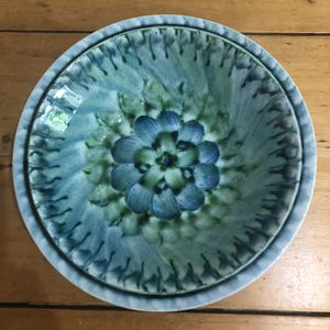 Puede incluir: Un cuenco de cerámica con un diseño floral en tonos azules y verdes. El cuenco tiene un centro elevado con un patrón floral y un borde texturizado. El cuenco se utiliza probablemente para servir o exhibir.