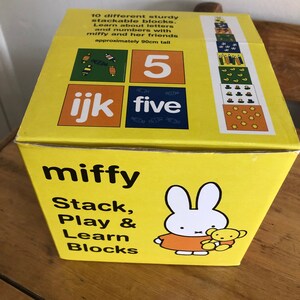 Puede incluir: Una caja amarilla de bloques apilables "miffy". La caja presenta el texto "Stack, Play & Learn Blocks" e ilustraciones del personaje Miffy. La caja también incluye el texto "10 bloques apilables resistentes diferentes" y mide aproximadamente 90 cm de alto.