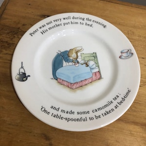 May include: White ceramic plate with a Peter Rabbit illustration. The plate has text around the rim, including "Peter was not very well during the evening." and "One table-spoonful to be taken at bed-time."