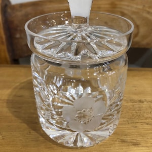 May include: A clear glass jar with a lid and a floral design. The jar has a textured surface and a flower etched into the side.