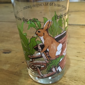 Puede incluir: Un vaso de vidrio transparente con una ilustración de un conejo marrón. El conejo está en una carretilla con plantas verdes. El texto en el vaso dice "Peter saw the gate... of Mr McGregor's garden."