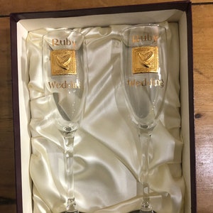 Könnte beinhalten: Zwei klare Champagnerflöten aus Glas mit goldener Aufschrift "Ruby Wedding". Die Flöten befinden sich in einer mit weißem Satin ausgekleideten Schachtel.