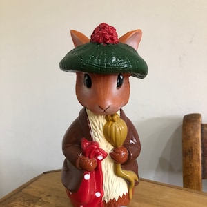 Puede incluir: Una figurita de conejo marrón que lleva un sombrero verde con un pompón rojo, una chaqueta marrón y una bufanda roja con lunares blancos. El conejo sostiene un palo amarillo.