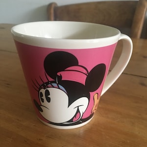 Peut inclure: Tasse en c&eacute;ramique rose avec un dessin de Minnie Mouse. L'int&eacute;rieur et la anse sont blancs. Le dessin de Minnie Mouse comprend des oreilles noires, un n&oelig;ud rose et un visage blanc. La tasse est pos&eacute;e sur une table en bois.